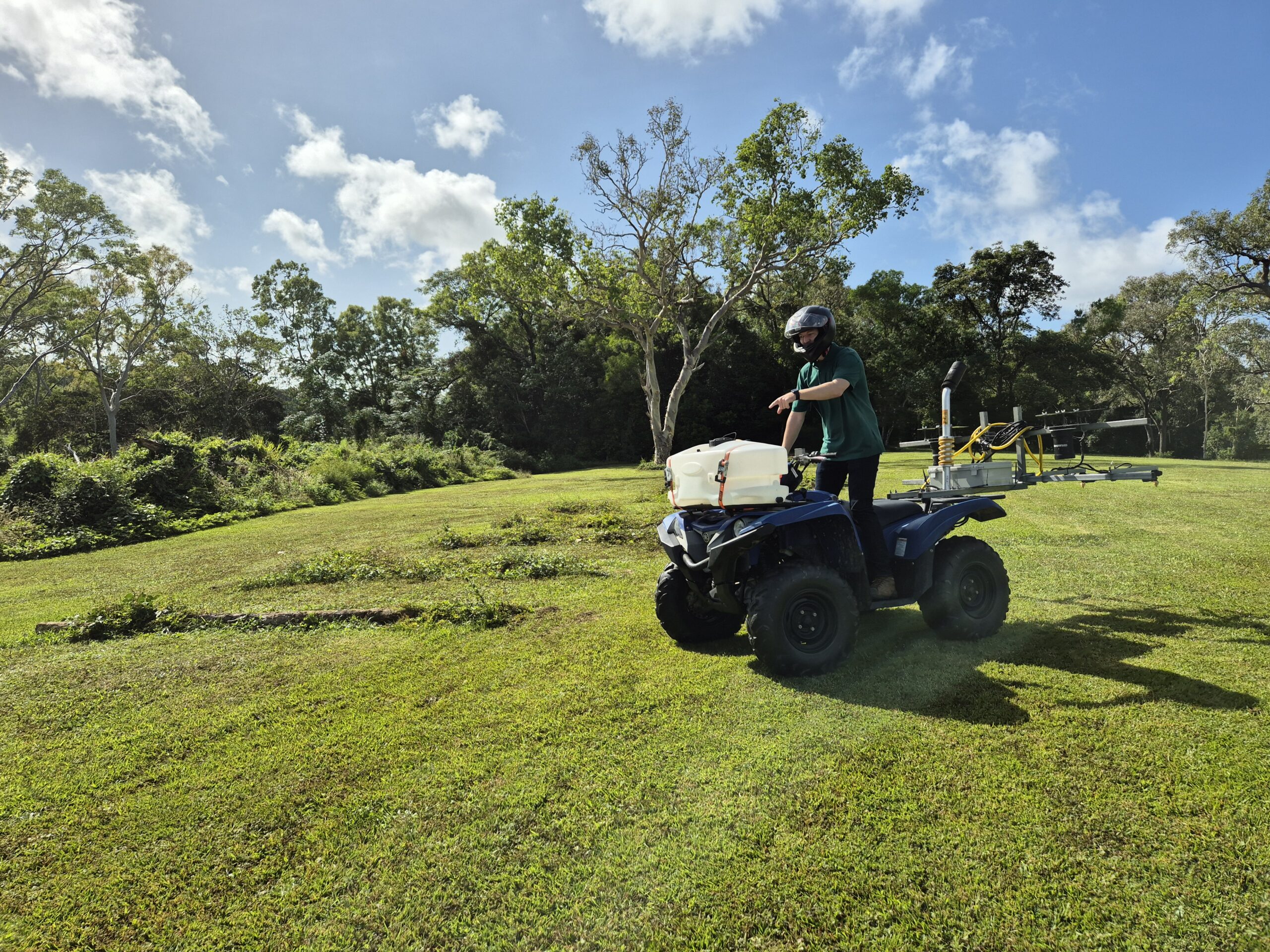 Man in a green field standing on a blue quad bike pointing to the ground in front of him.