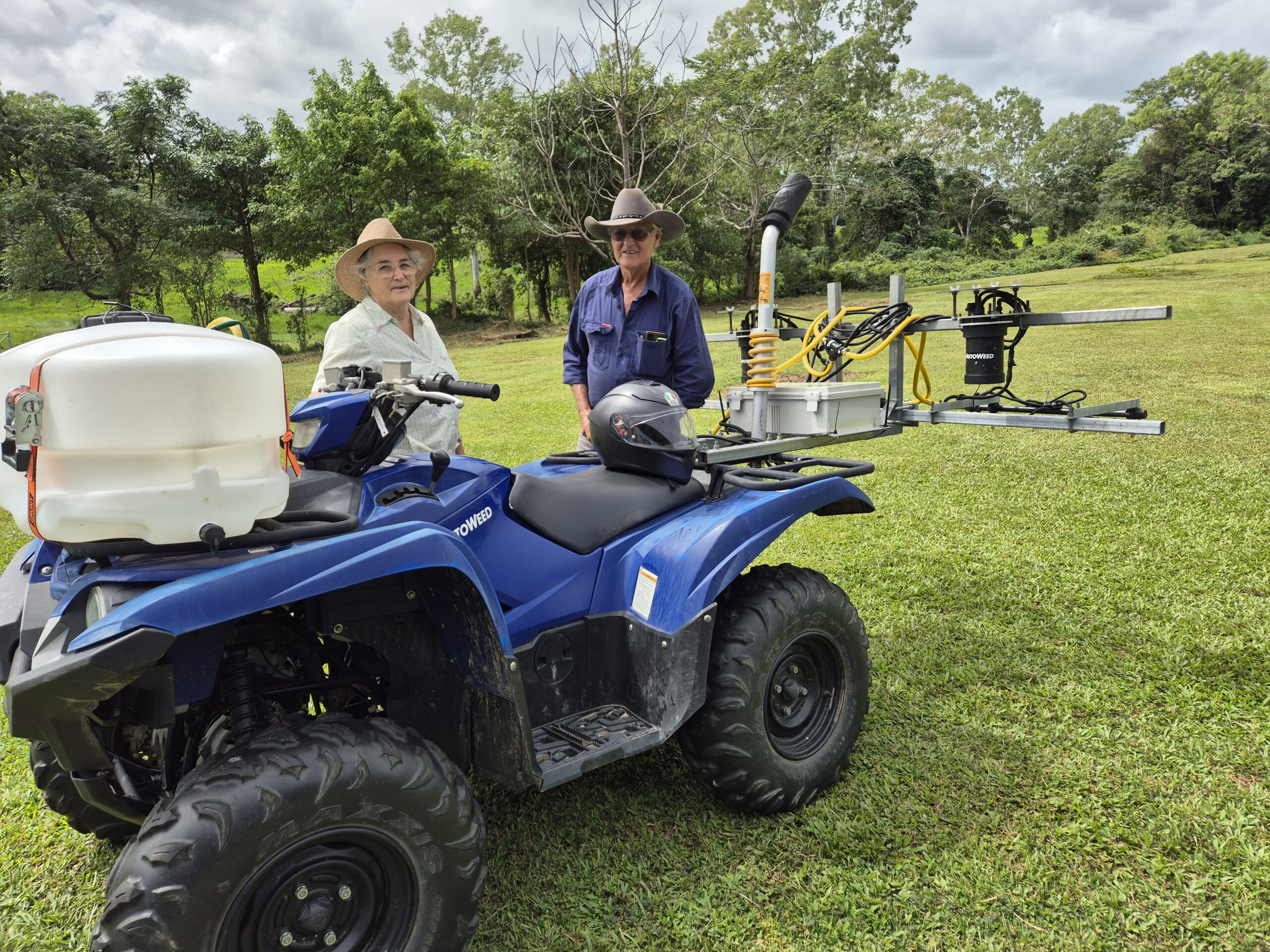 Blue quad bike with tank strapped to front and sensor at back. Judy and Peter standing behind in farm clothes and hats. 